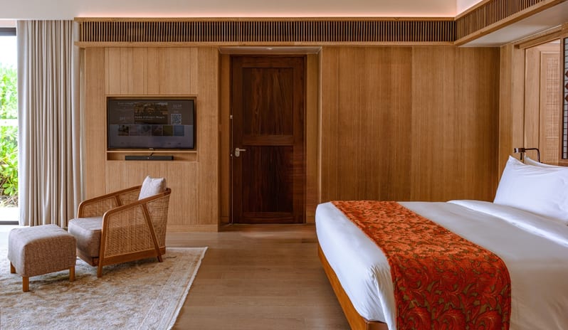 Sunset Water Villa with Private Pool light wood paneled walls, a king-sized bed with an orange patterned runner, a woven wicker armchair with an ottoman, and a recessed wall-mounted television at Kuda Villingili Resort Maldives by Kenwood Travel