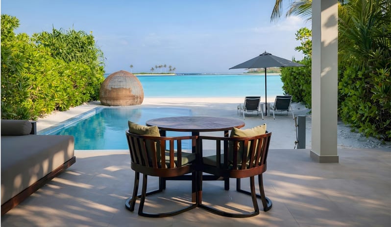 Sunset Beach Villa with Private Pool Outdoor patio of a luxury beach villa featuring a dark wood dining set, private infinity pool, and a wicker daybed pod on a white sand beach overlooking turquoise ocean water at Kuda Villingili Resort Maldives by Kenwood Travel