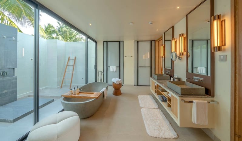 Sunset Water Villa with Private Pool grey stone freestanding bathtub, a double vanity with stone vessel sinks, and floor-to-ceiling glass doors leading to a private outdoor shower area with tropical greenery at Kuda Villingili Resort Maldives by Kenwood Travel