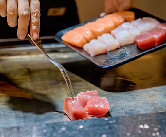 Teppanyaki by Mar-Umi Close-up of a chef using a fork to sear fresh tuna cubes on a flat-top teppanyaki grill, holding a platter of raw salmon and white fish in the background by Kenwood Travel