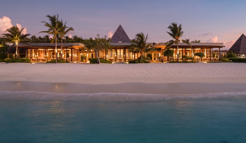 The Restaurant luxury tropical beachfront restaurant at dusk with illuminated pyramid-shaped roofs, palm trees, and white sand beach meeting turquoise ocean water at Kuda Villingili Resort Maldives by Kenwood Travel