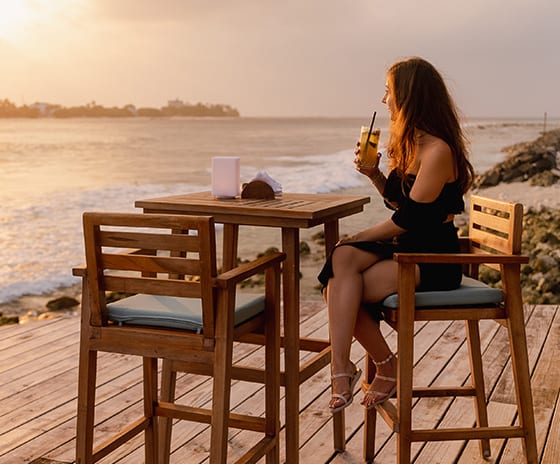 Raalhu Bar woman in a black dress enjoying a refreshing cocktail at a wooden high-top table on an oceanfront deck during a golden hour sunset in the Maldives at Kuda Villingili Resort Maldives by Kenwood Travel