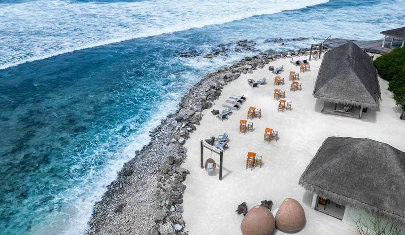 Aerial view of Kuda Villingili Resort Maldives featuring a white sand beach, turquoise ocean waves, and outdoor lounge seating with thatched-roof villas