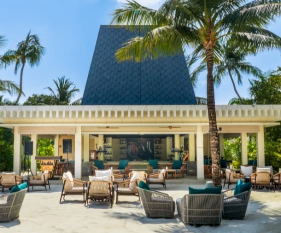 The Bar bar Exterior view of a luxury beach resort lounge with woven seating and turquoise pillows on a sandy patio, featuring a high-peaked dark roof pavilion and palm trees under a clear blue sky at Kuda Villingili Resort Maldives by Kenwood Travel