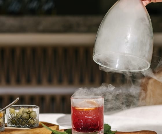 The Bar Close-up of a hand lifting a glass cloche to reveal a smoked red craft cocktail garnished with a dried citrus slice, served on a wooden board with a side of green olives at Kuda Villingili Resort Maldives by Kenwood Travel