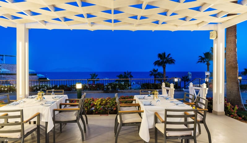 Adalar outdoor restaurant at twilight with glowing white pergolas, set dining tables, and tall palm trees against a blue evening sky at Royal Savoy Sharm El Sheikh by Kenwood Travel