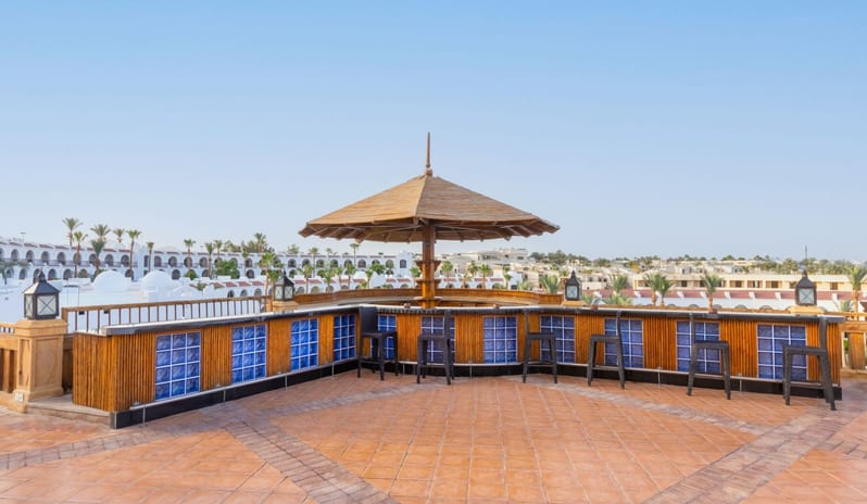 Le Balcony Rooftop bar at a luxury resort featuring bamboo paneling, blue glass accents, and a central umbrella over bar stools at Royal Savoy Sharm El Sheikh by Kenwood Travel