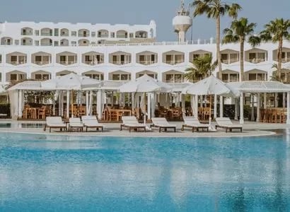 The Oasis Bar Bright poolside view of a modern white-walled tropical resort with palm trees and patio seating under white umbrellas at Baron Resort Sharm El Sheikh by Kenwood Travel