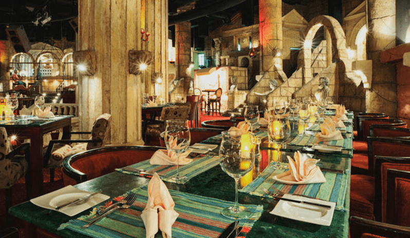 Caligula Hot Rock Long dining table set with wine glasses and folded napkins inside a luxurious ancient Roman themed restaurant with stone pillars at Royal Savoy Sharm El Sheikh by Kenwood Travel