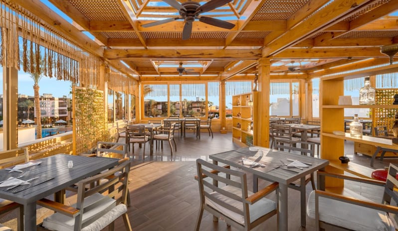 Tirana Bright and airy hotel terrace restaurant with a warm wood pavilion ceiling, set tables, and windows looking out onto a tropical resort landscape at Royal Savoy Sharm El Sheikh by Kenwood Travel