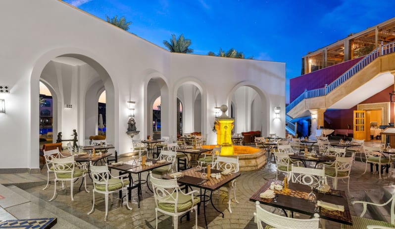 Rosmarino An elegant outdoor resort restaurant courtyard at twilight featuring white arches, a lit central fountain, and white patio dining tables at Royal Savoy Sharm El Sheikh by Kenwood Travel