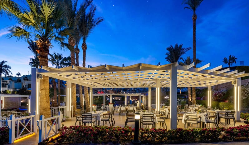 Adalar outdoor restaurant at twilight with glowing white pergolas, set dining tables, and tall palm trees against a blue evening sky at Royal Savoy Sharm El Sheikh by Kenwood Travel