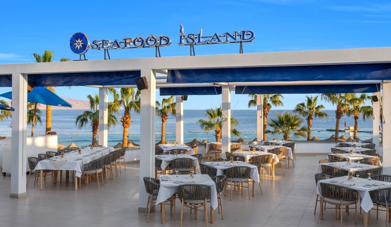 Seafood Island set tables, palm trees, and an ocean view under a clear blue sky at Royal Savoy Sharm El Sheikh by Kenwood Travel