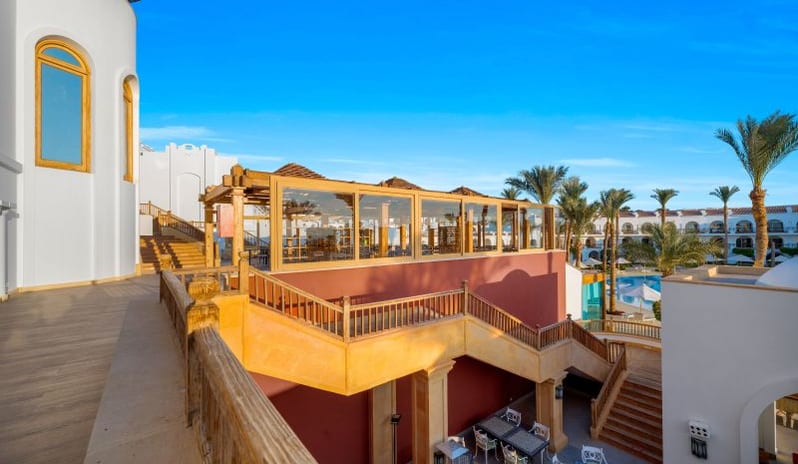 Tirana An elevated hotel patio with wooden railings and a glass-walled dining pavilion overlooking an outdoor restaurant courtyard and resort pool at Royal Savoy Sharm El Sheikh by Kenwood Travel