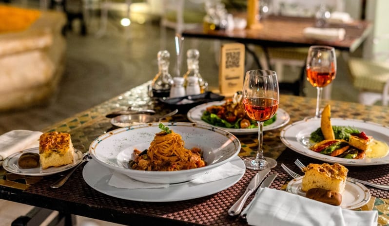 Rosmarino outdoor resort dinner setup showing a table arranged with fine dining pasta dishes, wine glasses, and condiments on a mosaic tile table at Royal Savoy Sharm El Sheikh by Kenwood Travel