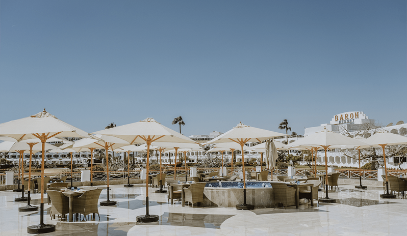 Sinai Restaurant beige umbrellas, wicker dining tables, and a central water fountain under a clear blue sky at Baron Resort Sharm El Sheikh  by Kenwood Travel