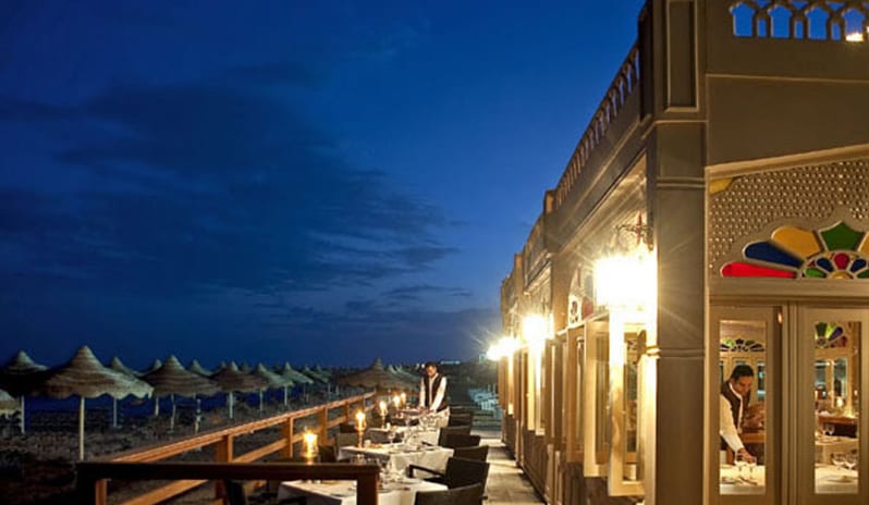 Taj Mahal RestaurantSide-angle view of an illuminated restaurant veranda at night, showcasing outdoor seating, a wooden railing, and a view of the dark ocean coastline at Baron Resort Sharm El Sheikh by Kenwood Travel