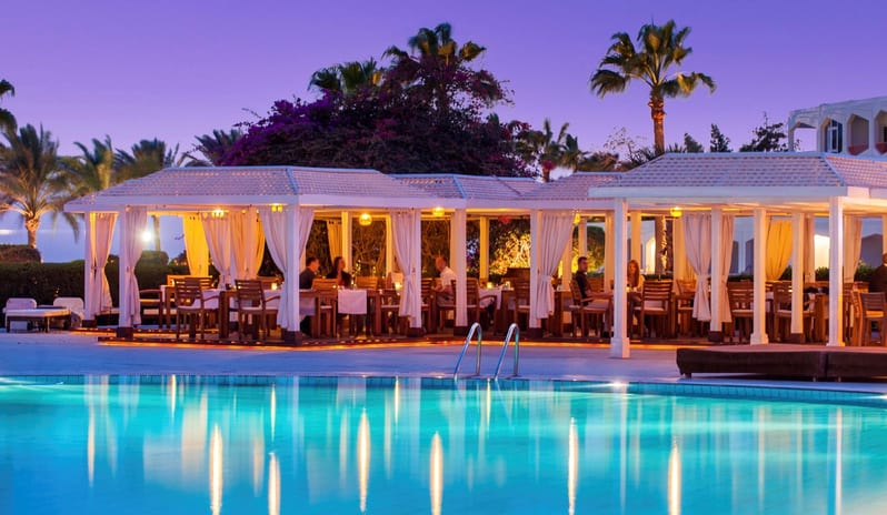 Oasis BBQ & Seafood Restaurant and Bar outdoor dining pavilions with white curtains next to a large resort pool under a vibrant purple sunset sky at Baron Resort Sharm El Sheikh by Kenwood Travel