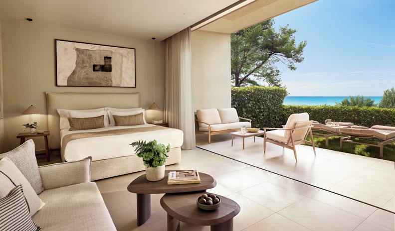 Deluxe Double Room Private Garden Sea View king-size bed, indoor sitting area, and large sliding doors that open seamlessly onto a private sun-drenched patio overlooking a calm blue ocean at Sani Club by Kenwood Travel