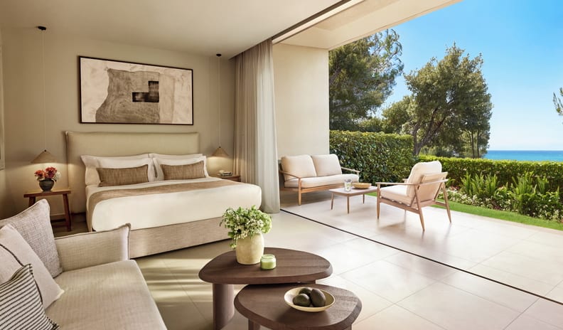 Double Room Ground Floor Sea View bedroom with an open-concept sliding wall leading to a private outdoor patio, featuring modern minimalist decor, neutral tones, and a view of the ocean and green garden at Sani Club by Kenwood Travel