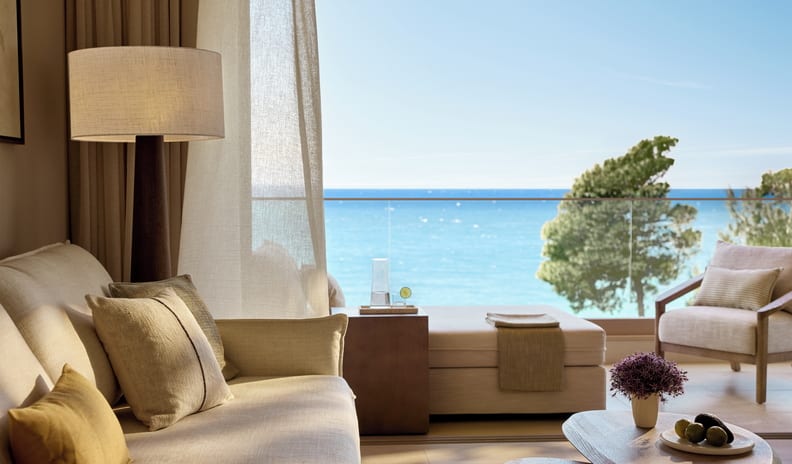 Family Suite Grand Balcony Sea View Luxury resort living room and outdoor terrace featuring neutral-toned linen sofas, a wooden coffee table with fresh fruit, and a sunbed overlooking a bright blue ocean at Sani Club by Kenwood Travel