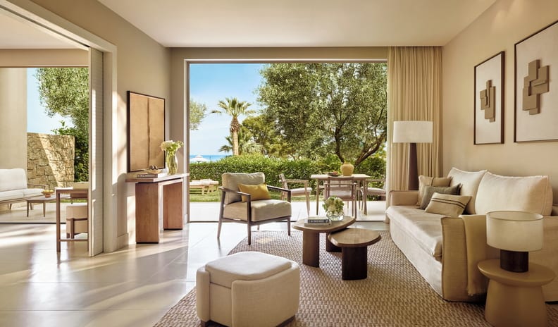 One Bedroom Bungalow Suite Private Garden Pool Front beige fabric sofa, nested wooden coffee tables, an accent armchair, and a glimpse into an adjacent bedroom through a wide doorway at Sani Club by Kenwood Travel