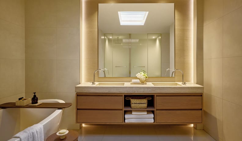 One Bedroom Bungalow Suite Private Garden Pool Front bathroom featuring a floating wooden double vanity with a beige stone countertop, dual undermount sinks with gooseneck faucets, a large square backlit mirror, and a partial view of a freestanding bathtub with a wooden tray at Sani Club by Kenwood Travel