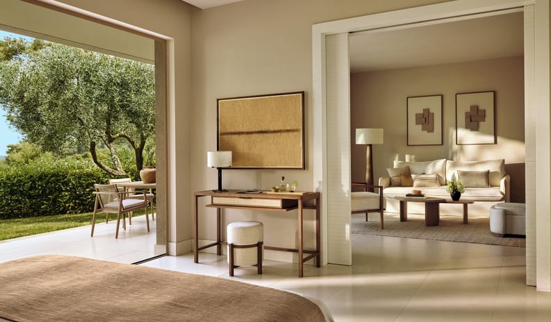 Two Bedroom Bungalow Suite Private Garden Sea View bedroom suite featuring a modern wooden vanity desk, a bed with a textured tan throw, and large glass doors opening to a sunny garden patio with olive trees at Sani Club by Kenwood Travel