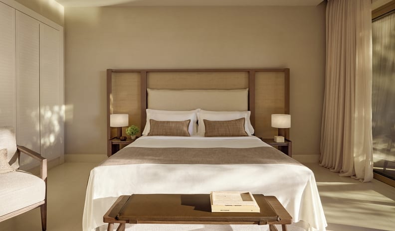 One Bedroom Bungalow Suite Private Pool Garden View bedroom with neutral linens, custom wooden headboard, bedside lamps, and a wooden bench in a bright, airy setting at Sani Club by Kenwood Travel
