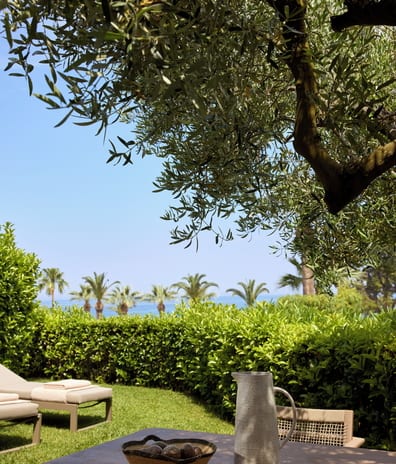 Two Bedroom Bungalow Suite Private Garden Sea View featuring a wooden dining table under an olive tree, looking out over a green lawn with sun loungers, hedges, and distant palm trees by the sea at Sani Club by Kenwood Travel