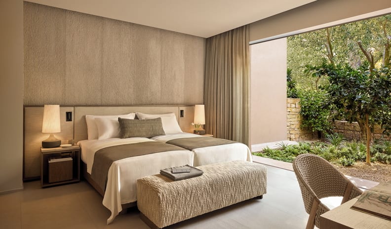 Three Bedroom Grand Residence Private Pool Garden View bedroom featuring neutral linens and a woven desk chair, looking out onto a sunny, private garden terrace with stone walls at Sani Asterias by Kenwood Travel