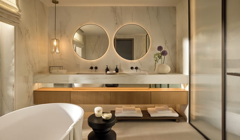 Three Bedroom Villa Pr. Pool Garden Viewbathroom of a luxury resort suite, featuring premium marble finishes, dual sinks, ambient pendant lighting, and crisp white folded towels on a slatted floor shelf at Sani Asterias by Kenwood Travel