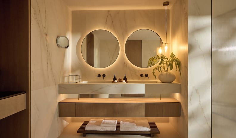 Three Bedroom Villa Pr. Pool Marina View luxury resort bathroom featuring a floating wooden vanity with an integrated marble trough sink, two round backlit LED mirrors, and warm ambient lighting against elegant veined marble walls at Sani Asterias by Kenwood Travel