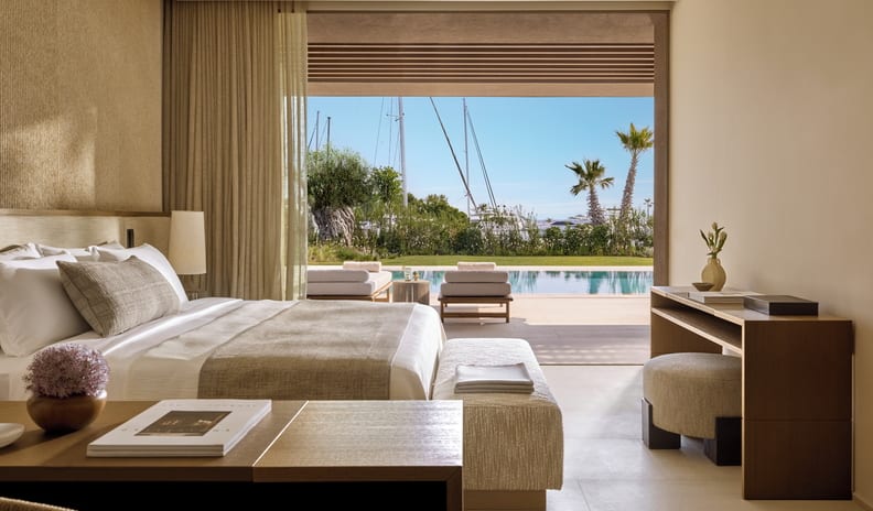 Three Bedroom Villa Pr. Pool Marina View Modern minimalist hotel suite with earth-toned decor, featuring a king bed that opens up to a private patio, swimming pool, and scenic marina view with palm trees at Sani Asterias  by Kenwood Travel