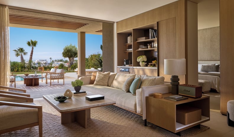 Three Bedroom Villa Pr. Pool Marina View luxury resort suite living room featuring a plush cream sofa, wooden coffee table, and seamless integrated wood wall cabinetry, opening completely to an outdoor poolside patio with marina views at Sani Asterias  by Kenwood Travel