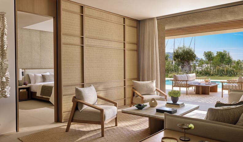 Three Bedroom Villa Pr. Pool Marina View living area with earth-toned decor and wooden armchairs, featuring a woven sliding partition wall opening into a bedroom, looking out onto a private outdoor patio with lounge seating and a swimming pool at Sani Asterias  by Kenwood Travel