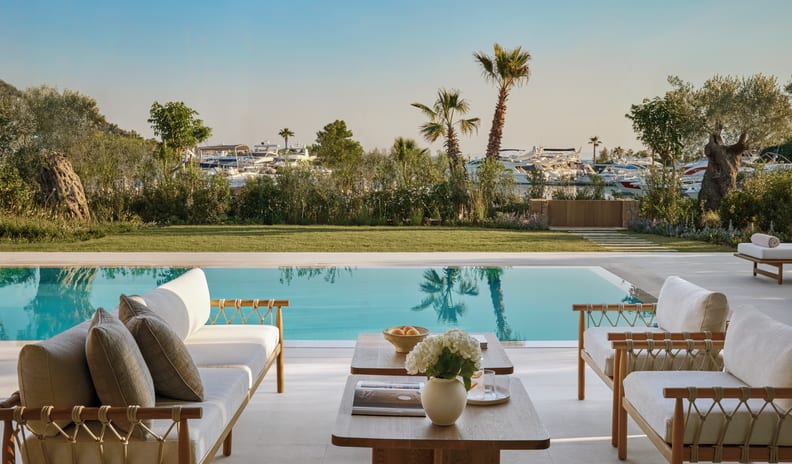 Three Bedroom Villa Pr. Pool Marina View lwooden sofas with woven rope details, a central coffee table with white hydrangeas, looking out across a private swimming pool toward a marina filled with luxury yachts and palm trees at Sani Asterias  by Kenwood Travel