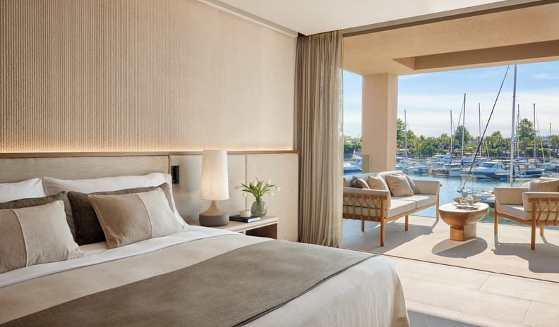One Bedroom Residence Grand Balcony Marina Front Elegant resort bedroom with a plush king bed and a bedside lamp, looking out toward a sunny harbor filled with boats and yachts at Sani Asterias by Kenwood Travel