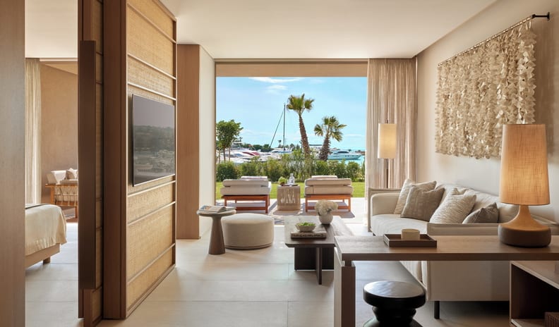 One Bedroom Residence Private Garden Marina Front Japandi-style hotel suite interior showcasing a seamless indoor-outdoor transition, a neutral color palette, textured wall installations, and a glimpse of an adjacent bedroom through a sliding wood partition at Sani Asterias by Kenwood Travel