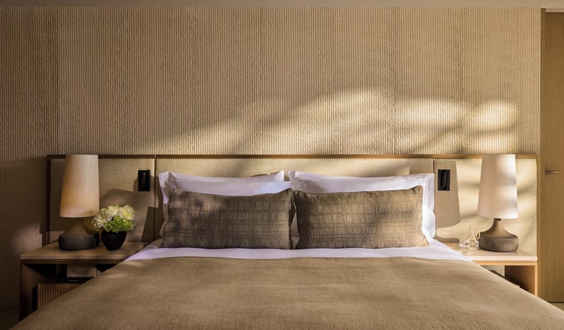 One Bedroom Residence Private Garden Marina Front bedroom setup with a light wood headboard, sand-colored textured wallpaper, elegant bedside lighting, and a small flower bouquet on the nightstand at Sani Asterias by Kenwood Travel