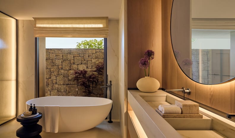 One Bedroom Residence Private Garden Marina Front bathroom design with Japandi influences, showcasing warm wood paneling, a sleek integrated sink with stone finishes, purple allium flowers in a ceramic vase, and a black accent side table next to a deep soaking tub at Sani Asterias by Kenwood Travel