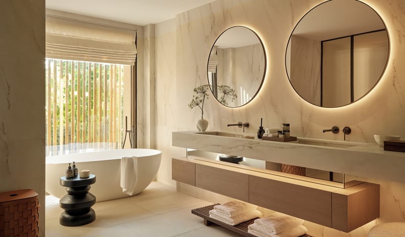 Sani Asterias Two Bedroom Grand Residence Balcony Beach Front SV bathroom featuring a modern freestanding soaking tub by a window, a marble double vanity with floating wooden cabinets, and two large round backlit mirrors against a marble wall-Kenwood Travel