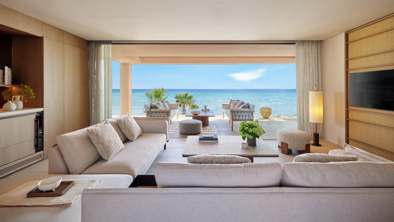 Two Bedroom Grand Residence Balcony Beach Front SV living room with contemporary grey sofas, light wood paneling, and a wide open-air glass wall leading to a private beach patio with outdoor seating overlooking the seaat Sani Asterias by Kenwood Travel