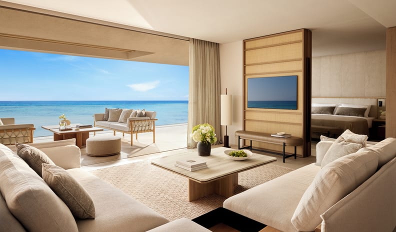 Two Bedroom Grand Residence Balcony Beach Front SV open-plan living room with cream sofas, a flat-screen TV mounted on a woven partition wall, a partial view of the bedroom, and a wide glass opening to a beachfront balcony terrace at Sani Asterias by Kenwood Travel