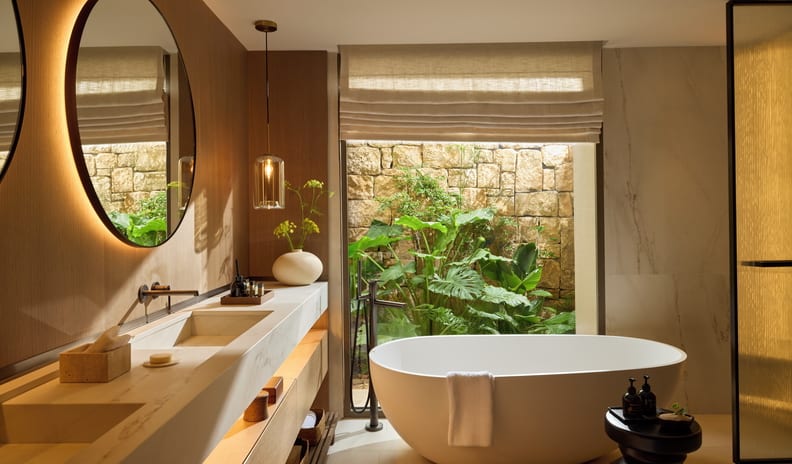 Two Bedroom Residence Pr. Pool Beach Front SV bathroom with a double stone vanity, round backlit mirrors, warm wood paneling, and a Roman shade window at Sani Asterias by Kenwood Travel