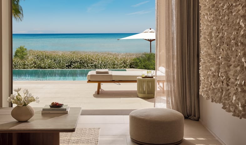 Two Bedroom Residence Pr. Pool Beach Front SV living room featuring a round fabric ottoman, sheer curtains, and textured wall art overlooking a patio pool at Sani Asterias by Kenwood Travel