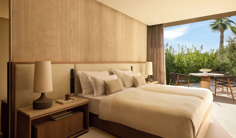 Two Bedroom Grand Residence Pr. Pool Marina Front bedroom featuring a textured beige accent wall, neutral bedding, and large sliding doors opening to a sun-drenched private patio with outdoor seating and a palm tree at Sani Asterias by Kenwood Travel