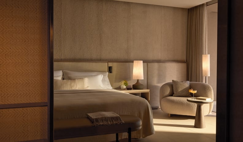 Two Bedroom Grand Residence Pr. Pool Marina Front bedroom at dusk, featuring a neutral bed setup, a textured ribbed accent wall, and a cozy bouclé armchair with a small side table holding a drink under glowing ambient lamp lighting at Sani Asterias by Kenwood Travel