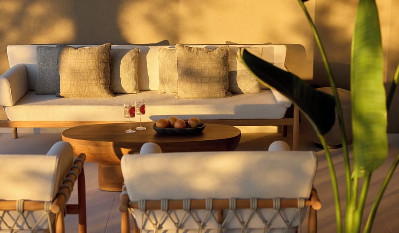 Two Bedroom Grand Residence Pr. Pool Marina Front cream-colored minimalist sofa with textured pillows, two matching armchairs, a solid wood coffee table with drinks, and a large tropical plant in the foreground under warm sunset shadows at Sani Asterias by Kenwood Travel