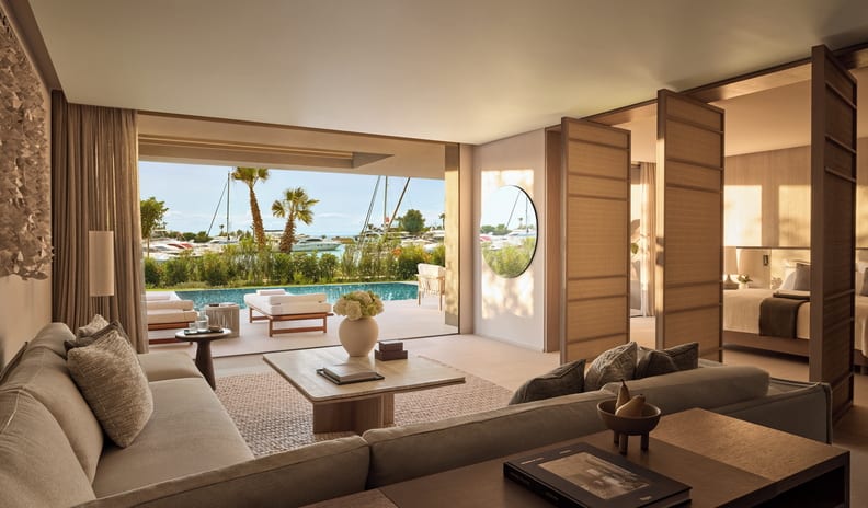 Two Bedroom Grand Residence Pr. Pool Marina Front open-plan living room with beige sofas, sliding screen partitions leading into a bedroom, and large open doors revealing a private pool terrace with sun loungers overlooking a marina with yachts and palm trees at Sani Asterias by Kenwood Travel